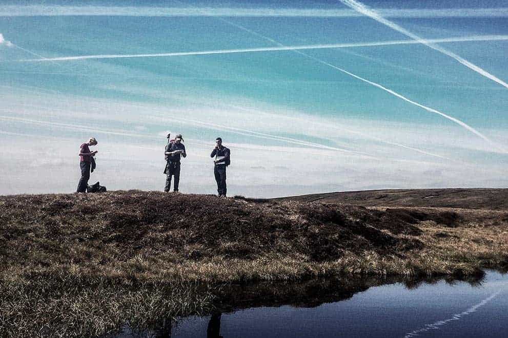Three people stand by a small moorland pond whilst practicing navigation on a Hill and Moorland Leader Award training course