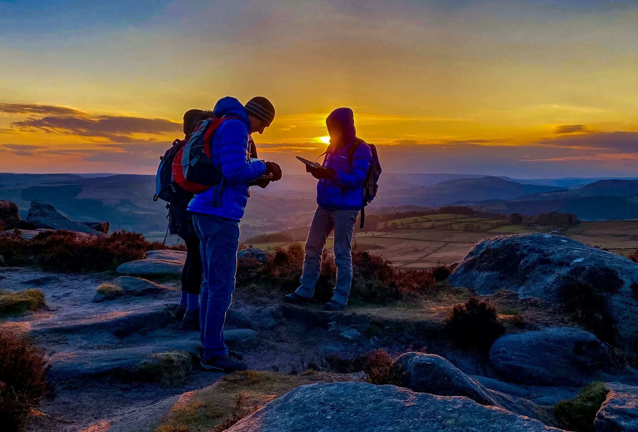 A group learn intermediate navigation using a map and compass as the sun sets whilst on a navigation course in the peak District.
