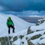 A winter mountaineer stands on top of a snowy ridge in Scotland with two ice axes in his hands.