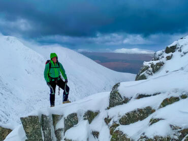 A winter mountaineer stands on top of a snowy ridge in Scotland with two ice axes in his hands.