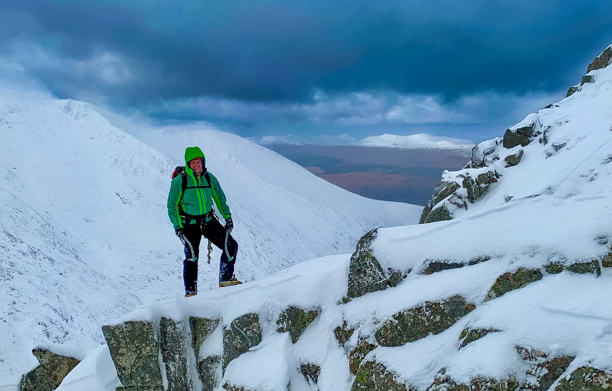 A winter mountaineer stands on top of a snowy ridge in Scotland with two ice axes in his hands.