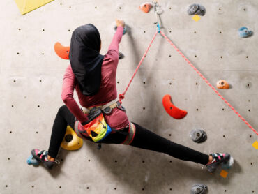 A female muslim climber leads a climb on an indoor wall for her Climbing Wall Development Instructor.