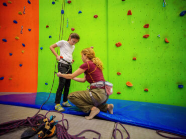 A climbing instructor helping a child with a climbing harness at a climbing wall.