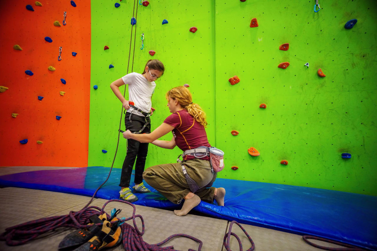 A climbing instructor helping a child with a climbing harness at a climbing wall.