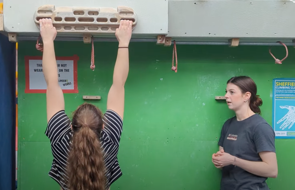 a woman hanging on a beastmaker climbing hangboard whilst chatting with a friend