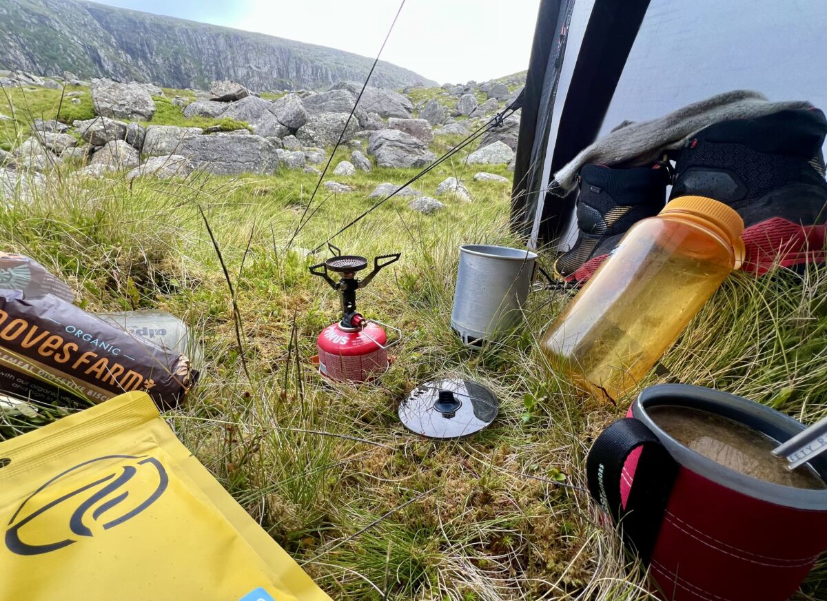 A view from a tent on a wild camp in the Welsh mountains. A gas camping stove, a mug with soup, biscuits and a water bottle.