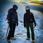 Two ladies stand on Cairngorm in evening light carrying ice axes and wearing crampons whilst on a winter mountaineering course in Scotland.