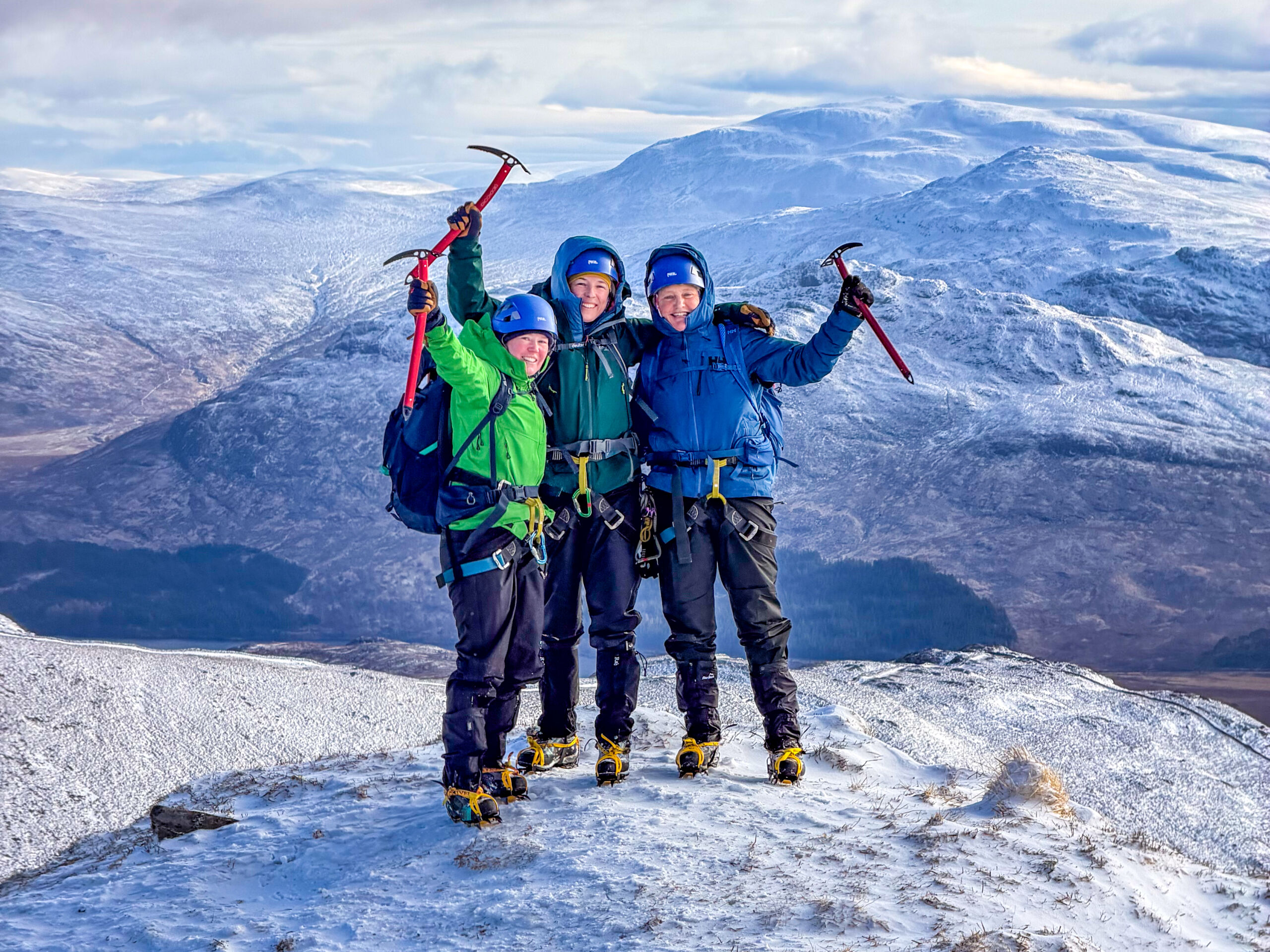 Three ladies on a winter skills course in Scotland, stand triumphantly holding ice axes on the summit of the East ridge of Beinn a Chairoann.